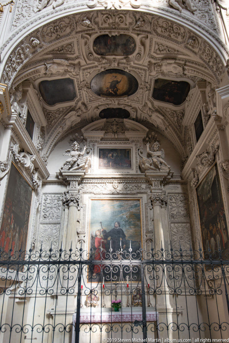 Ornate side chapel with central altarpiece, white stucco relief, multiple paintings, painted vault, and iron screen.