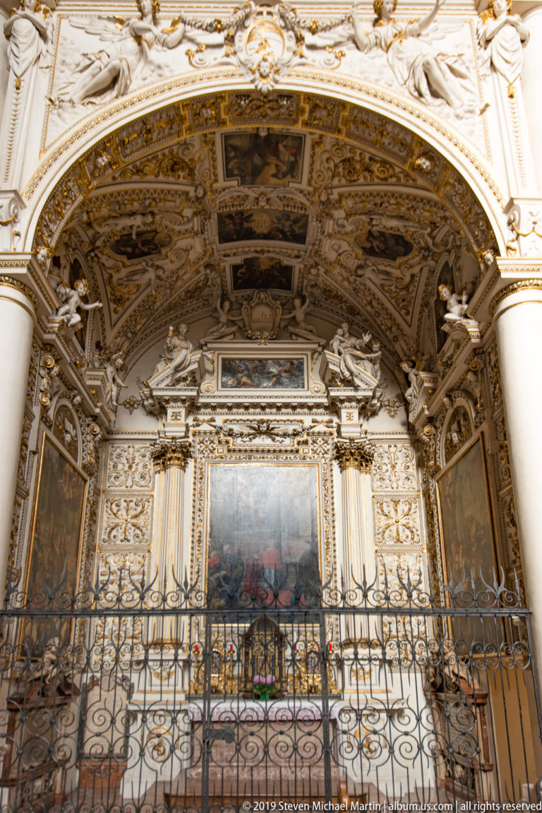 White stucco side chapel with large central painting, sculpted figures, decorated ceiling panels, and a wrought-iron barrier.