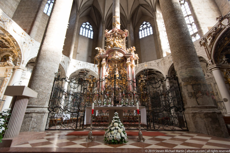 Closer frontal view of the ornate high altar with pink columns, gilded sculpture, iron screen, and tall Gothic windows behind.