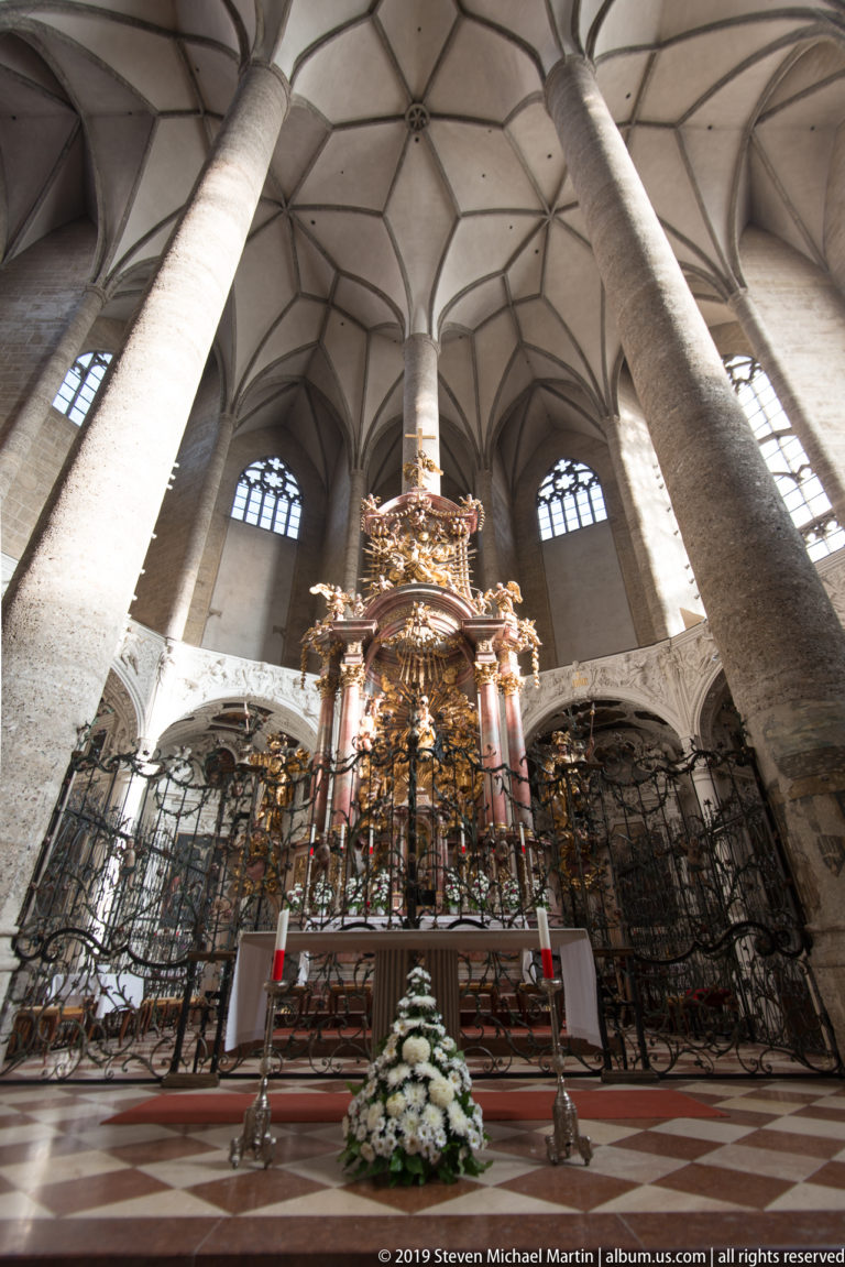 Frontal view of an ornate gilded high altar behind ironwork, framed by massive stone columns and a patterned church floor.