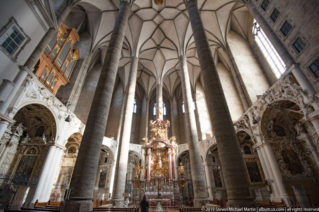 Wide interior view with tall stone columns, rib vaults, high windows, ornate side chapels, and a richly decorated central altar.