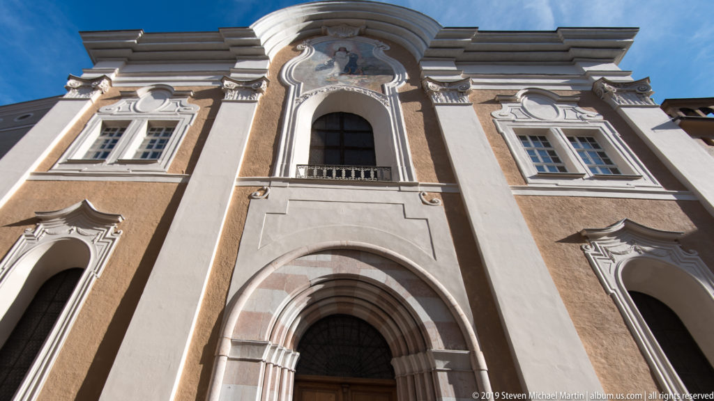 Low-angle view of the Franziskanerkirche façade with arched doorway, tall central window, white stucco trim, and sculpted pediment.