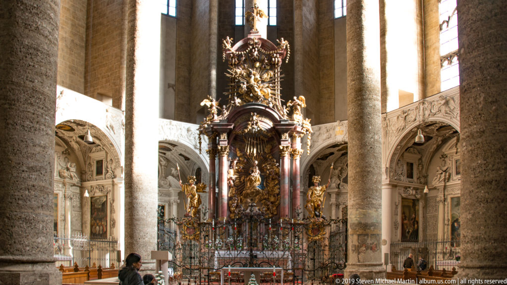 Ornate high altar illuminated by strong side light between tall stone columns, with a visitor standing to the left.