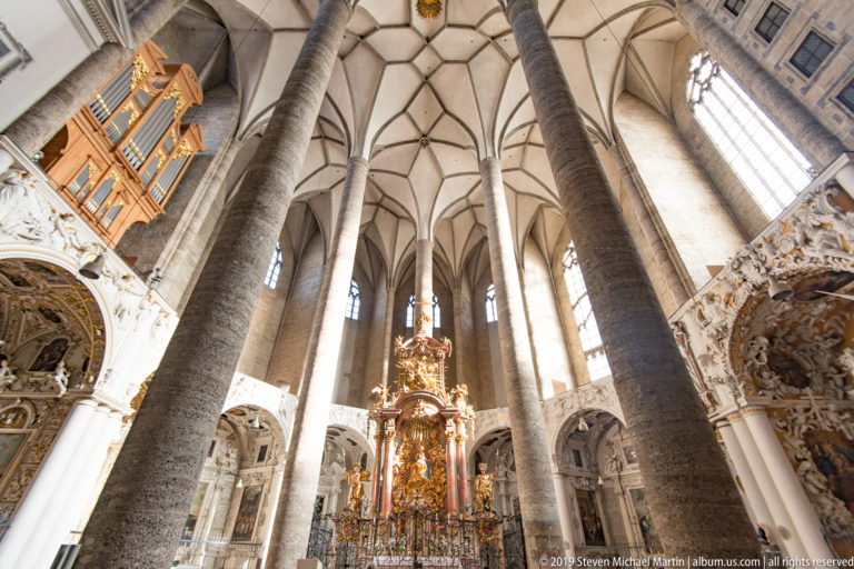 Interior of Salzburg’s Franziskanerkirche with tall Gothic columns and ribbed vaults leading toward a gilded Baroque high altar.