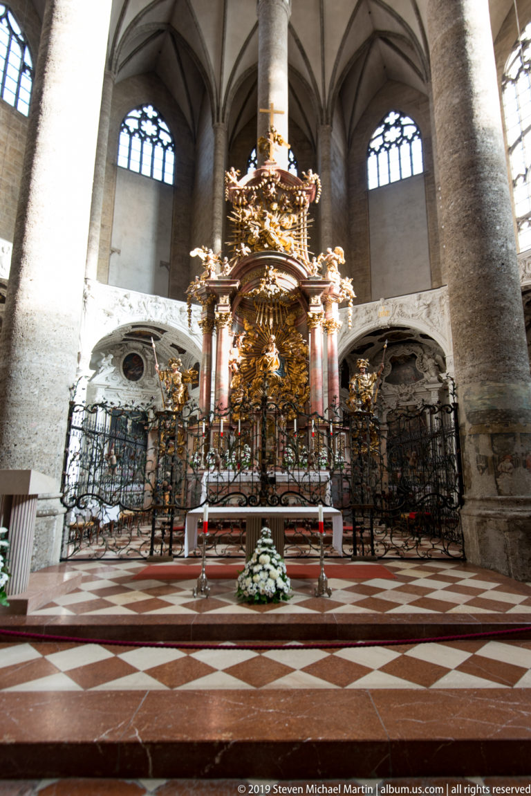 High altar centered between massive Gothic columns above a red-and-white patterned floor, with ironwork and flowers in front.