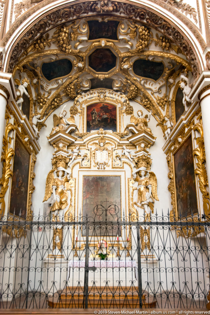 Highly gilded side chapel with ornate altar, religious paintings, sculpted decoration, painted ceiling panels, and iron screen.