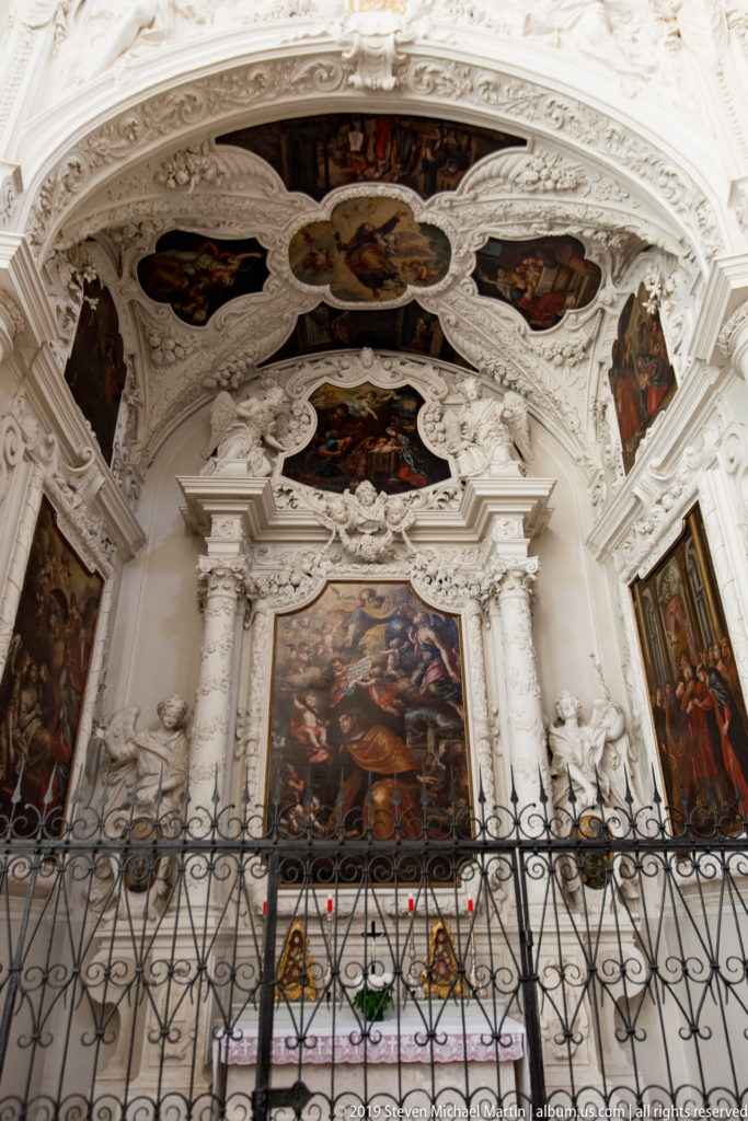 Baroque side chapel with large central painting, paired white columns, sculpted figures, painted vault panels, and iron screen.