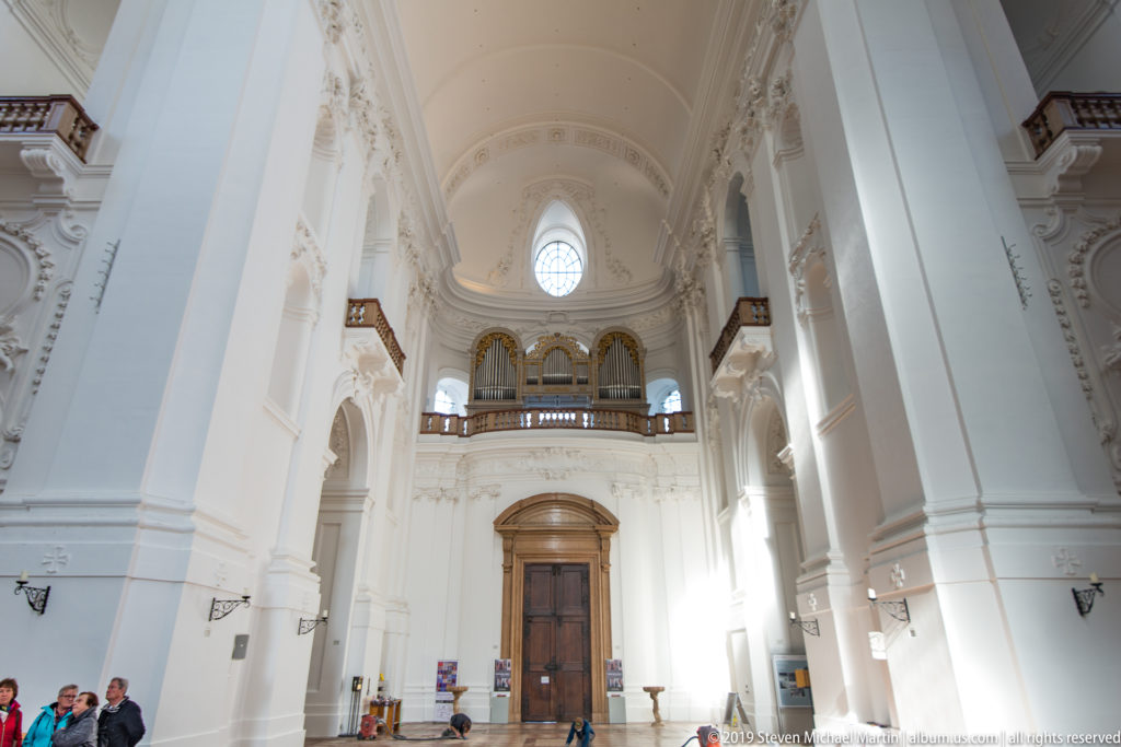 Tall white nave of the Kollegienkirche looking toward the organ gallery, entrance door, and high round window.
