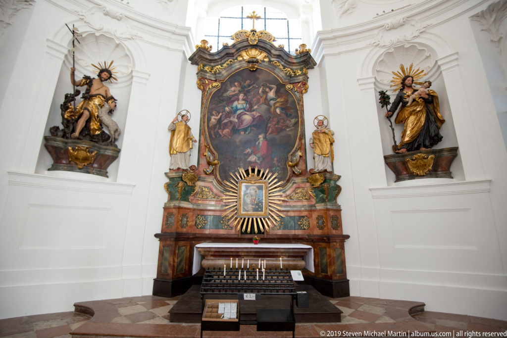 Ornate side altar with a large painted altarpiece, gilded decoration, and sculpted saints against white church walls.