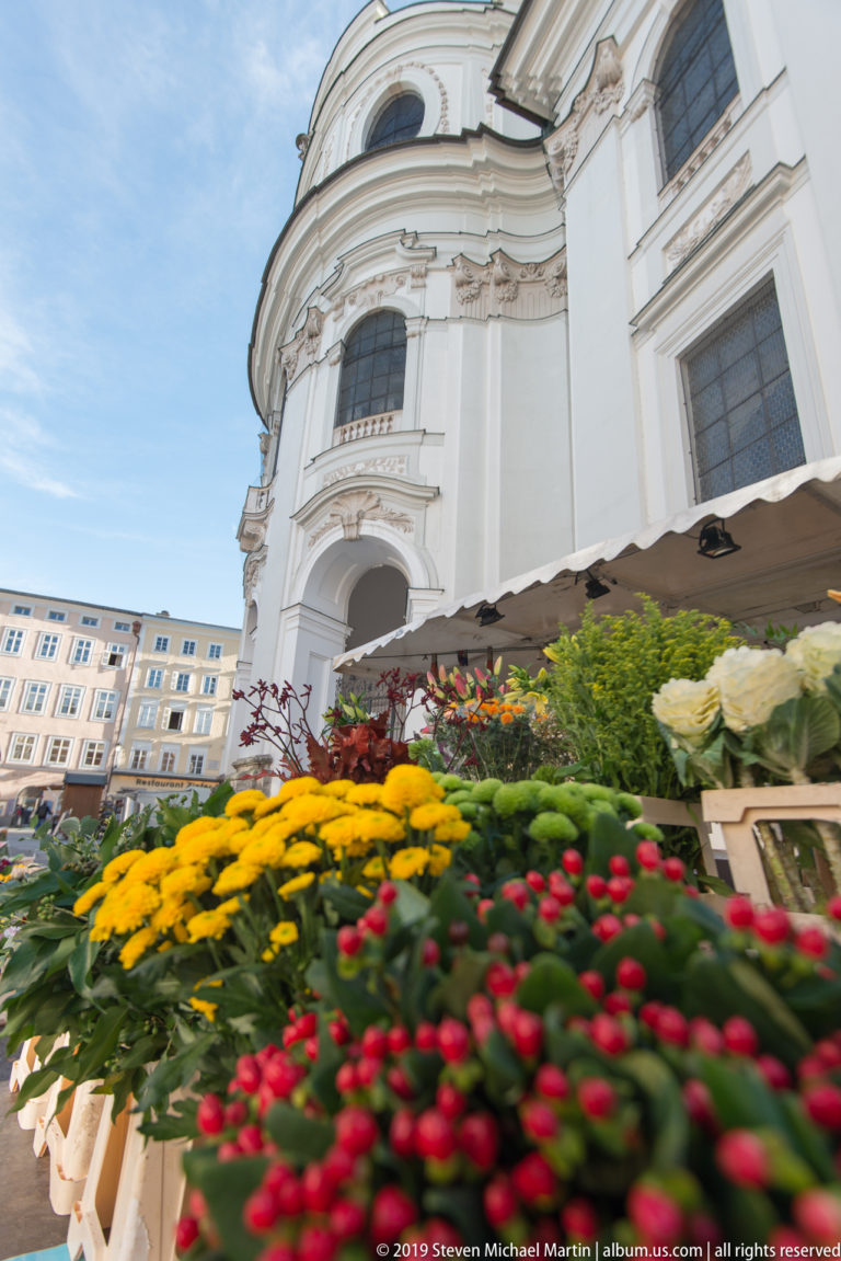 Colorful flower market display in front of the white Baroque façade of Salzburg’s Kollegienkirche.