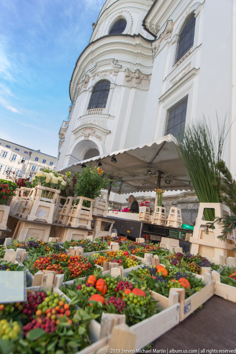 Fruit and vegetable market stalls beneath the curved white Baroque exterior of the Kollegienkirche in Salzburg.