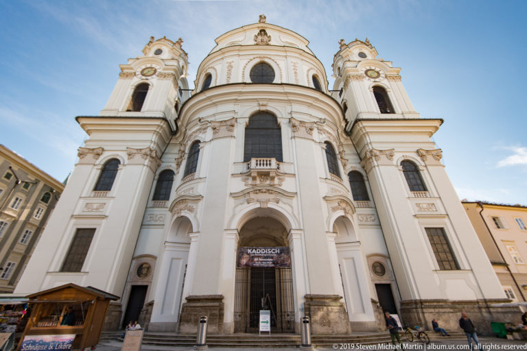 Low-angle view of Salzburg’s white Baroque Kollegienkirche with rounded central façade, tall windows, and twin towers.