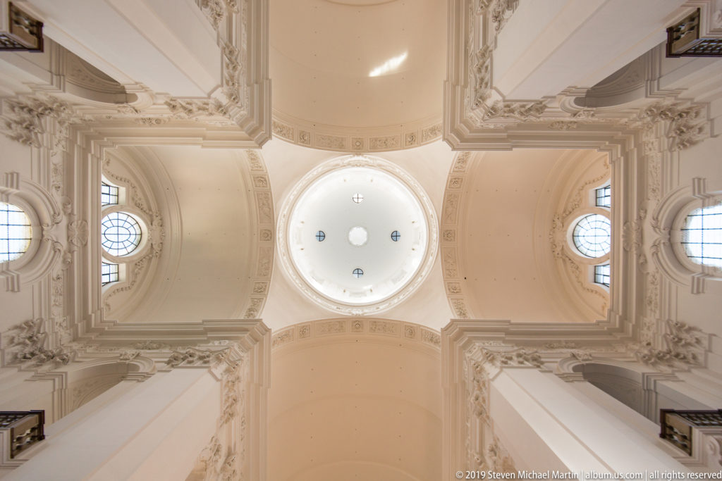 Upward symmetrical view of pale Baroque vaults, molded cornices, round windows, and a bright central opening.