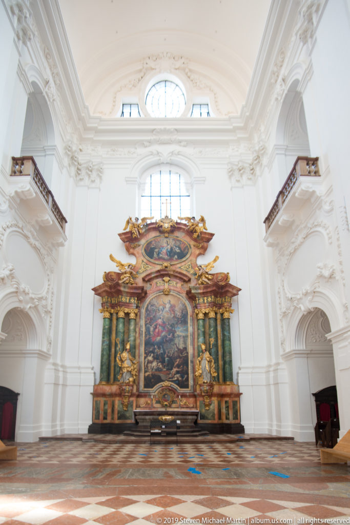 Wide interior view across patterned stone flooring to a colorful side altar framed by white arches and high windows.