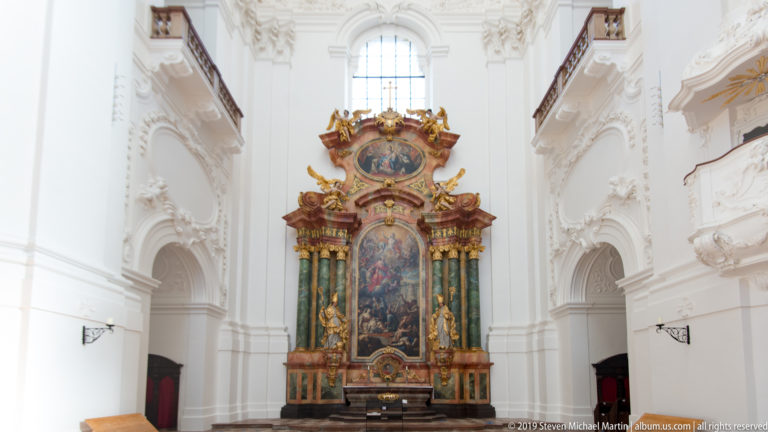Polychrome Baroque side altar with green columns and gilding centered between tall white arches in the Kollegienkirche.