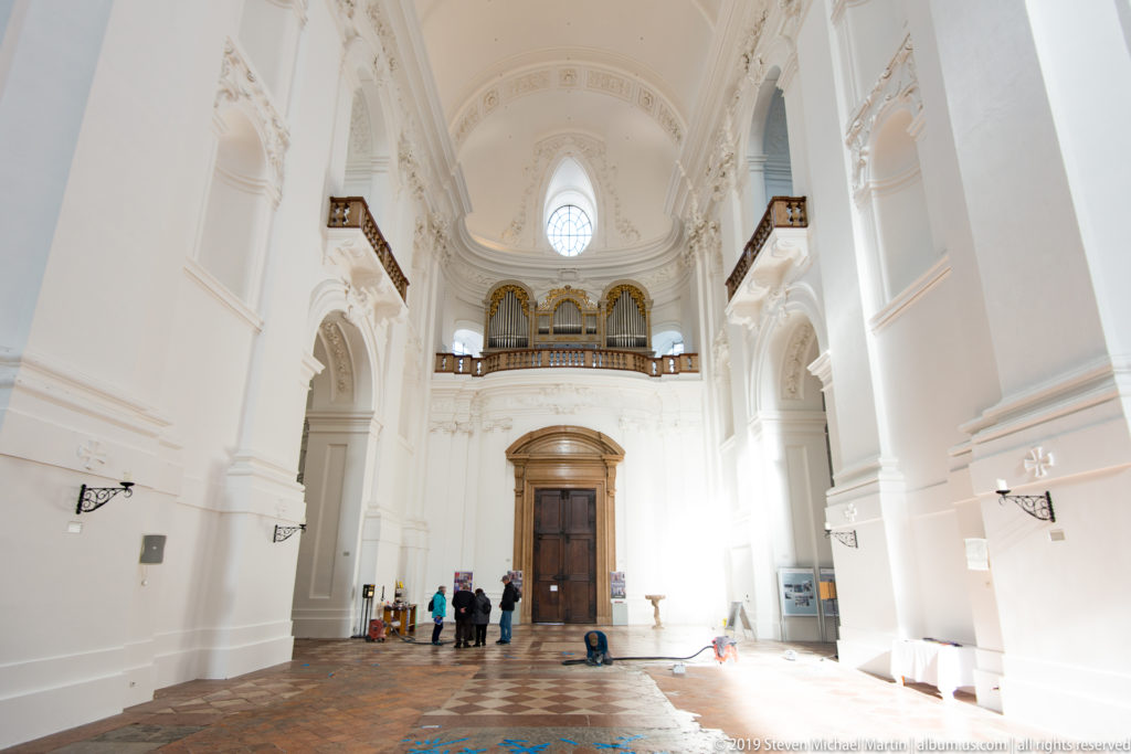 Wide white nave with patterned stone floor, tall arches, visitors, and the organ gallery above the entrance.