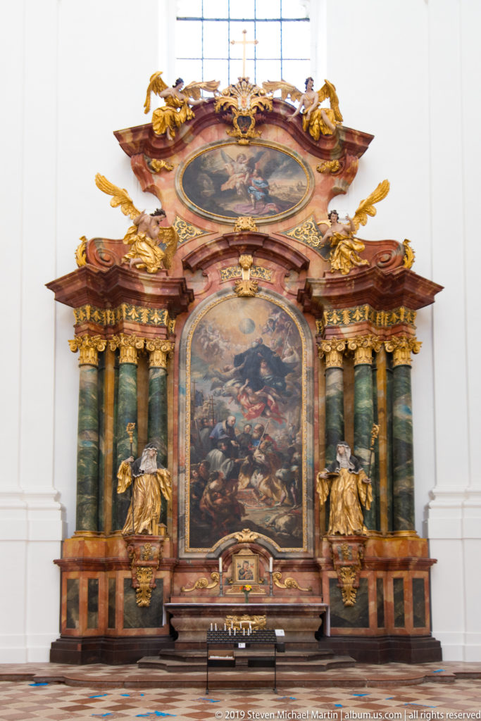 Large Baroque side altar with green columns, gilded figures, and two painted scenes against a white wall.