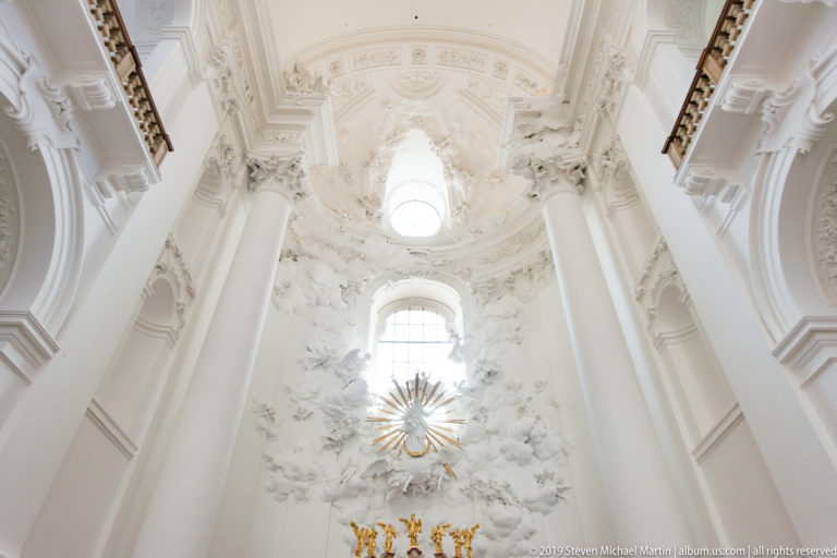 Upward view of bright windows, white sculptural stucco, and a gilded radiating emblem in the Kollegienkirche.