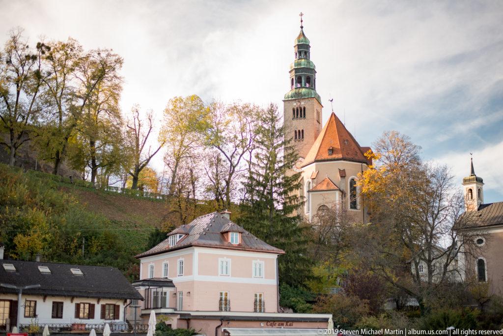 Wide hillside view of Pfarre Mülln with square green-topped tower, red roof, autumn trees, and a smaller tower at right.