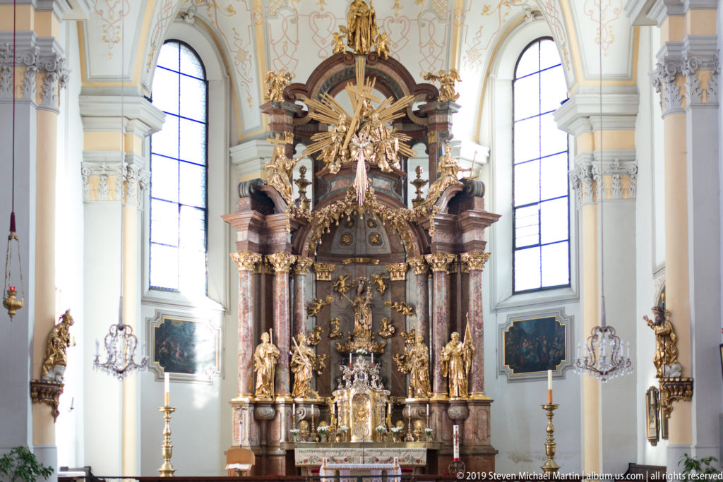 Frontal view of Pfarre Mülln high altar with marble columns, gilded statues, tall windows, candles, and side paintings.