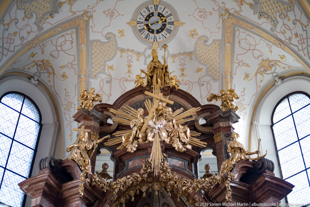 Upper section of the Baroque high altar with gilded rays and sculpture beneath a clock, flanked by tall arched windows.