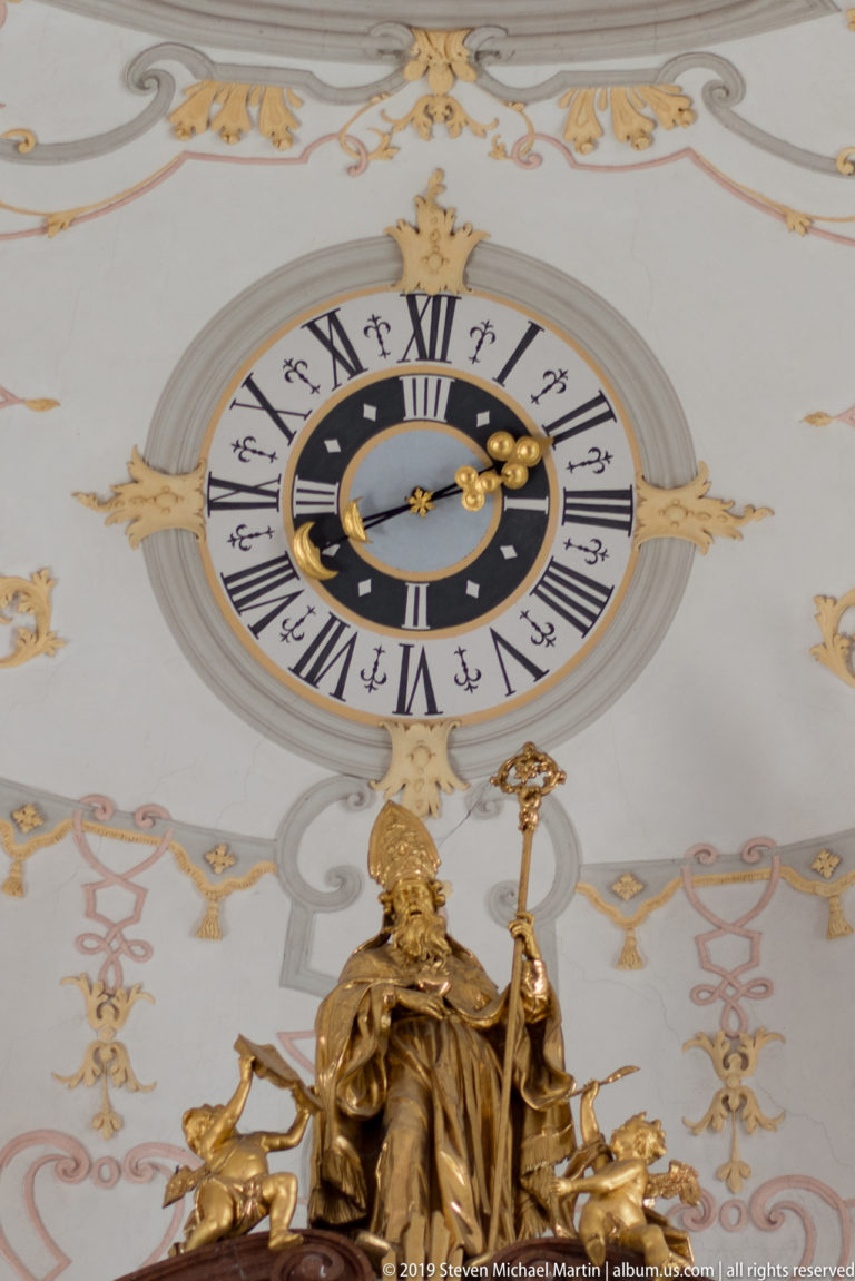 Roman-numeral clock framed by pale and gold ceiling decoration above a gilded figure at the altar crest.