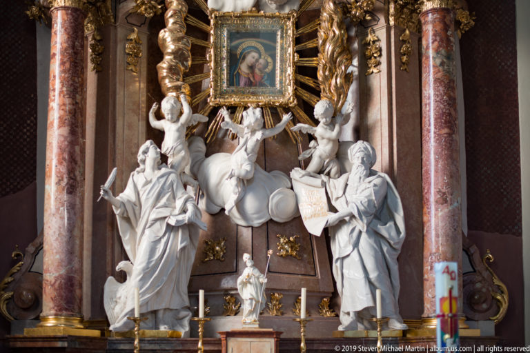 Frontal view of a Marian side altar with Virgin-and-Child image, white statues, cherubs, gilded rays, and columns.