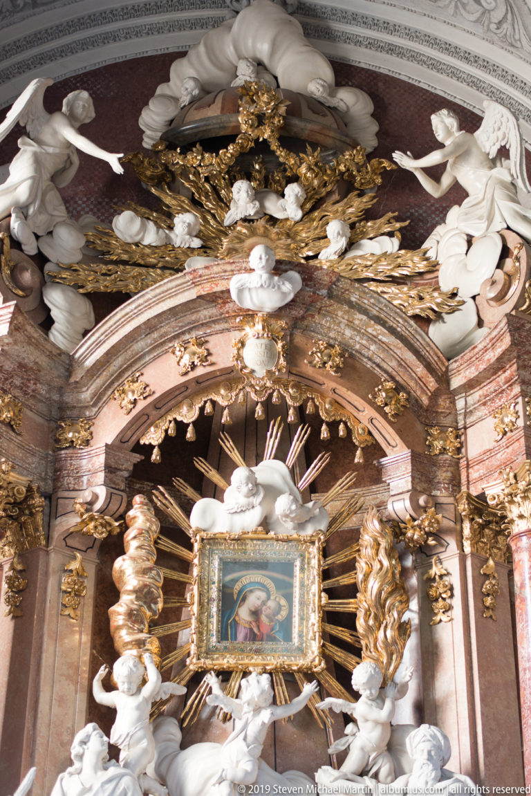 Ornate side altar with white sculpted saints and angels surrounding a small Marian image beneath gilded rays and canopy work.