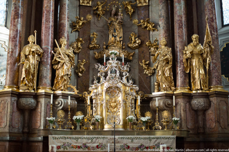 Lower section of an ornate Baroque high altar with gilded saints, marble columns, candles, and a bright central tabernacle.