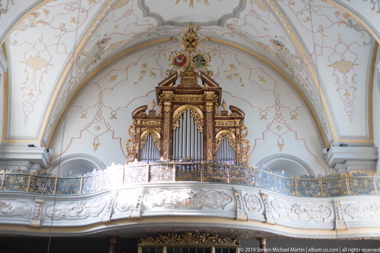 Large gilded pipe organ centered on a curved gallery beneath a decorated white vaulted ceiling.