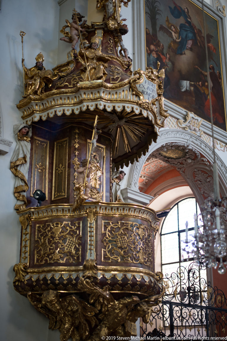 Ornate gilded Baroque pulpit with dark panels and carved canopy beside an arched church window.