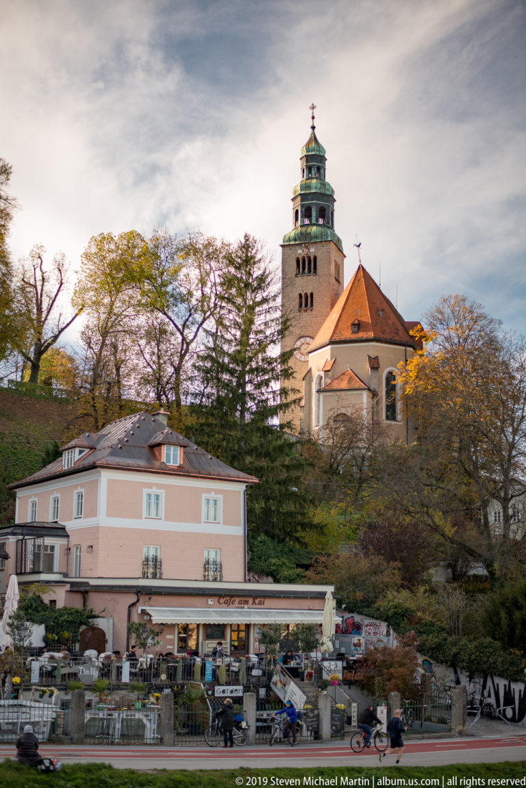 Mülln Parish Church rising above a pink building, café terraces, and autumn trees, with its green-crowned clock tower prominent.