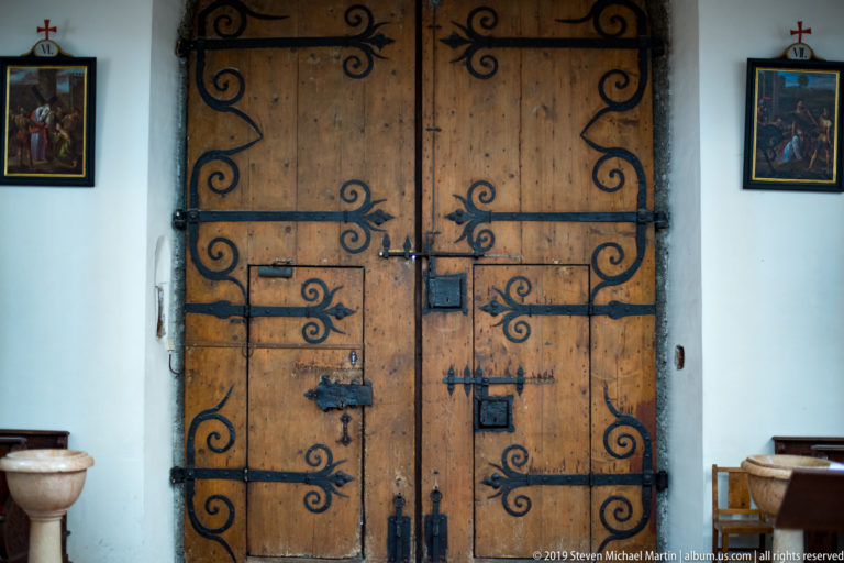 Closed double timber church doors with symmetrical black wrought-iron strapwork, flanked by small religious pictures and stone basins.