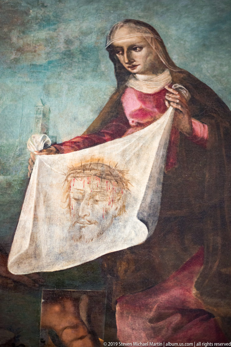 Painted Saint Veronica holding a white veil marked with the face of Christ against a blue-green background.