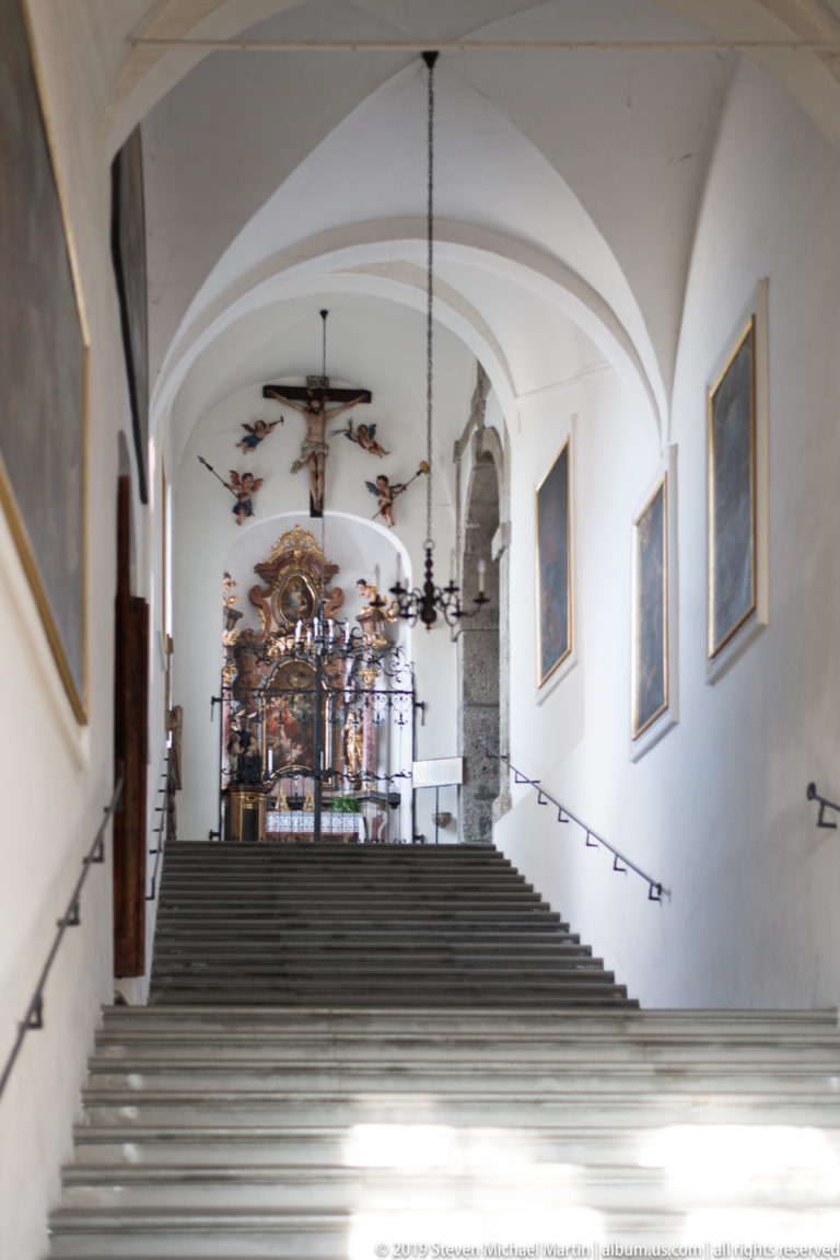 Wide stone staircase inside Pfarre Mülln rising beneath white vaults toward an ornate altar area and hanging crucifix.