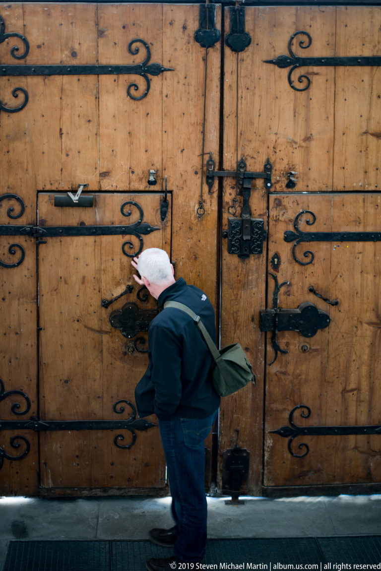 Visitor beside a massive timber church door covered with black wrought-iron hinges, straps, scrolls, and locks.