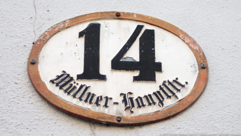 Oval enamel plaque reading “14 Müllner Hauptstr.” in black lettering on a white wall.