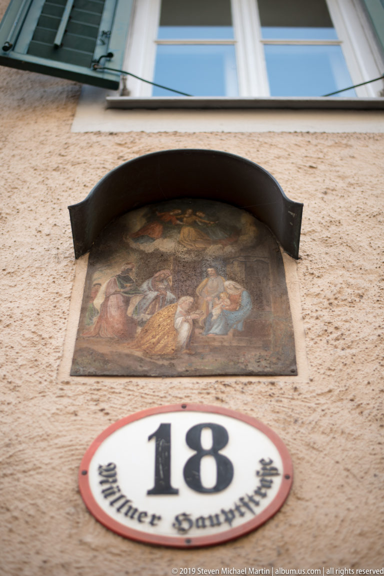 Weathered religious wall painting above an oval “18 Müllner Hauptstr.” plaque on a pale stucco façade.