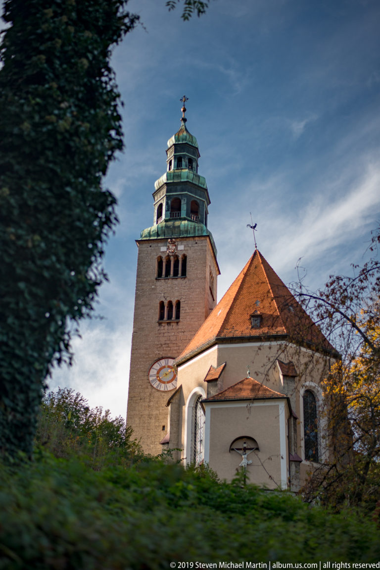 Mülln Parish Church with square clock tower, green tiered crown, red roof, and grassy slope beneath.