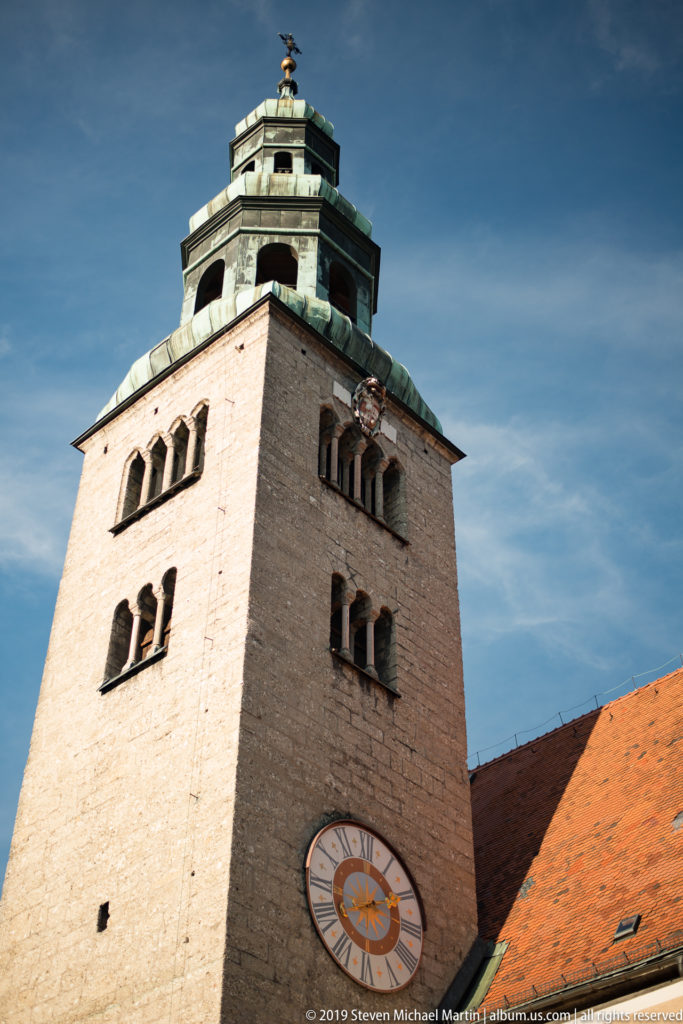 Low-angle view of Pfarre Mülln square tower with arched openings, clock face, red roof, and green-patinated lantern.