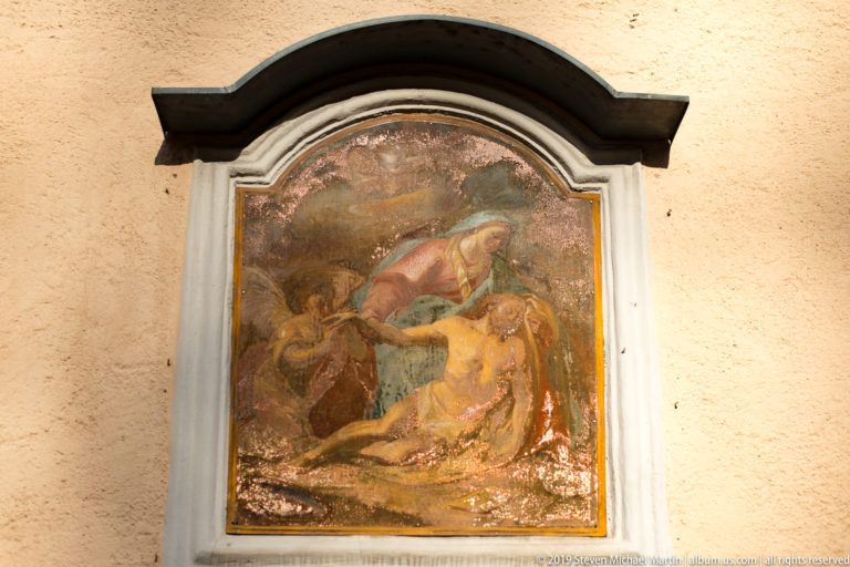 Weathered framed religious mural with figures on an ochre exterior wall beneath a curved protective hood.