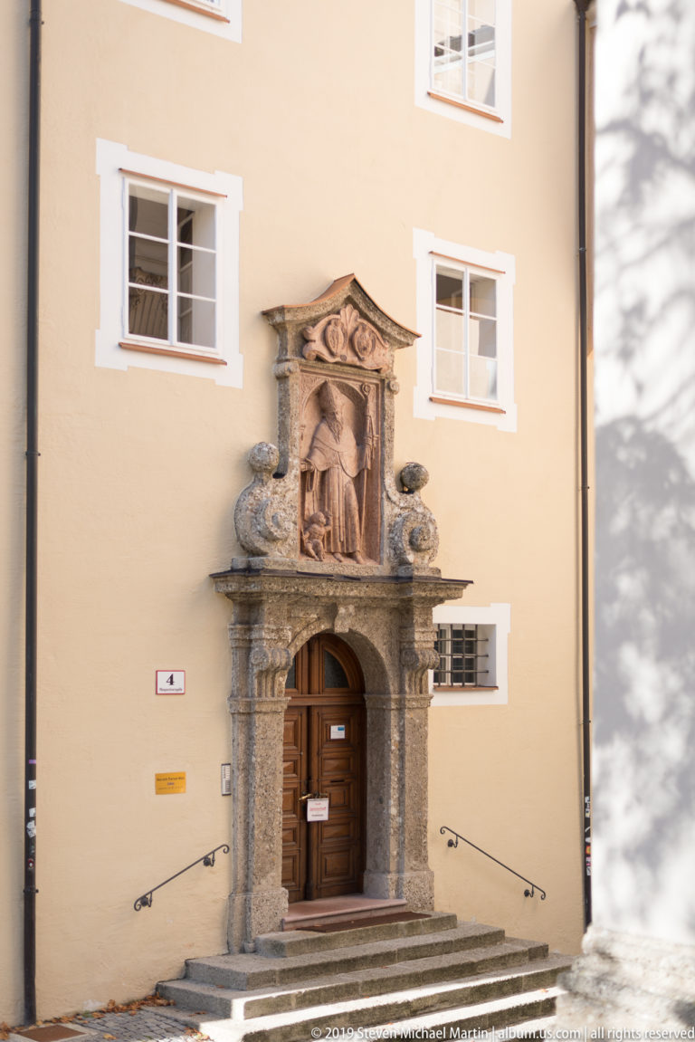 Stone church portal with wooden door, steps, heavy moldings, and sculpted relief set in an ochre façade.