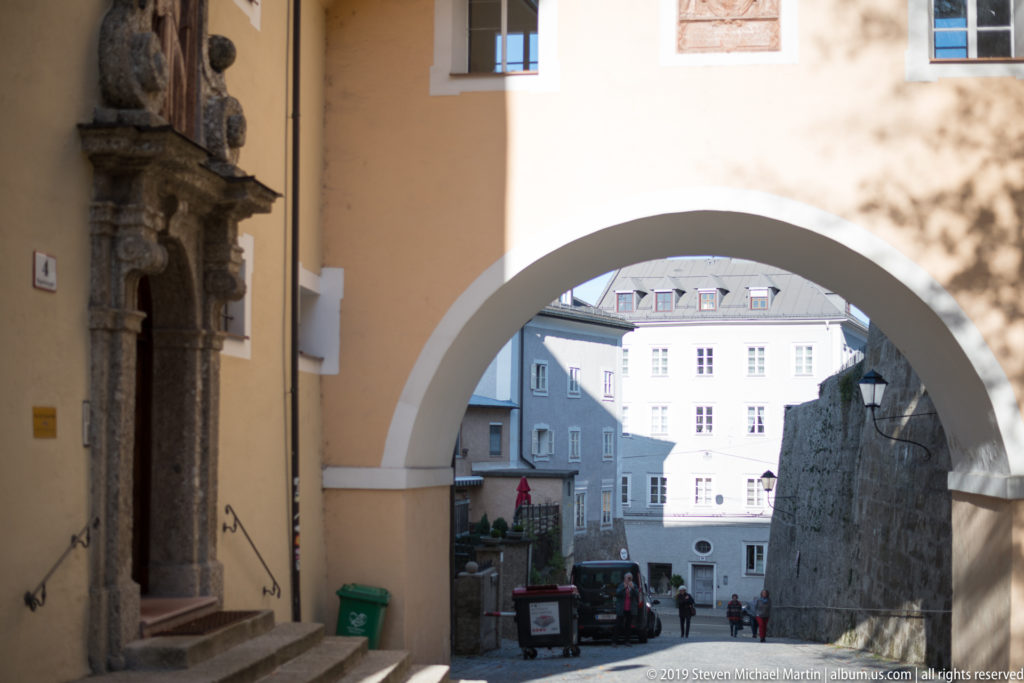 Large stucco archway beside Pfarre Mülln framing pedestrians, pale town buildings, lamps, and a stone wall beyond.