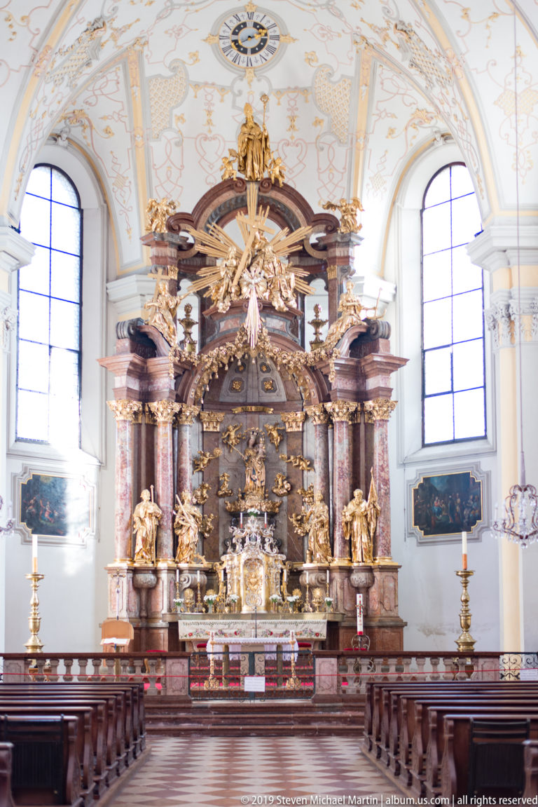 Wide nave view with wooden pews leading toward the ornate high altar beneath a decorated white vault.