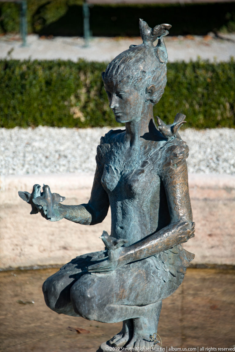 Blue-green bronze sculpture of a seated stylized woman with small birds perched on her head, shoulder, and outstretched hand.