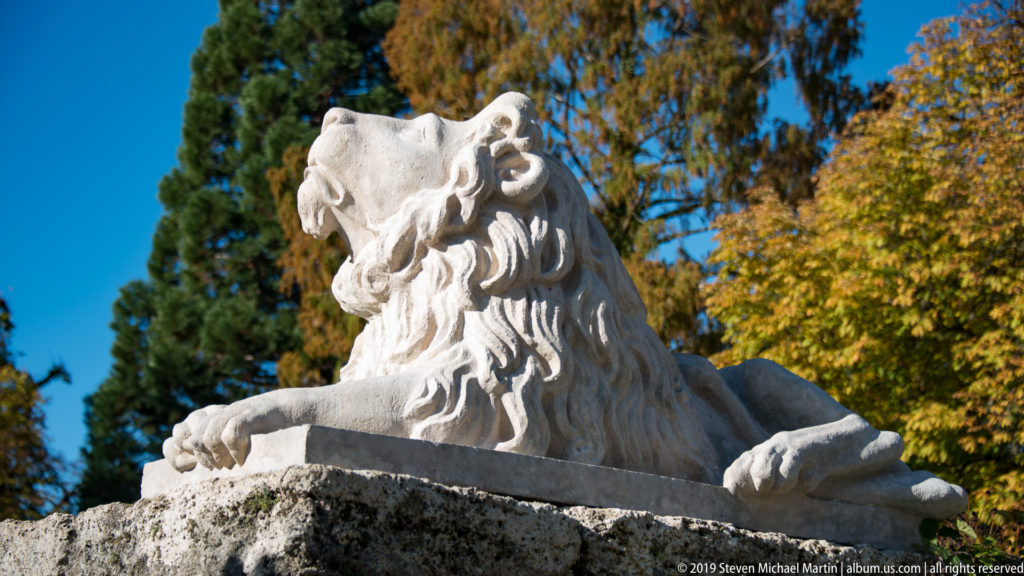 Reclining pale stone lion with deeply carved mane on a low plinth against blue sky and autumn trees.