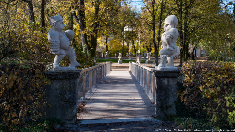 Straight garden walkway and narrow bridge flanked by two stone dwarf sculptures among clipped hedges and autumn trees.