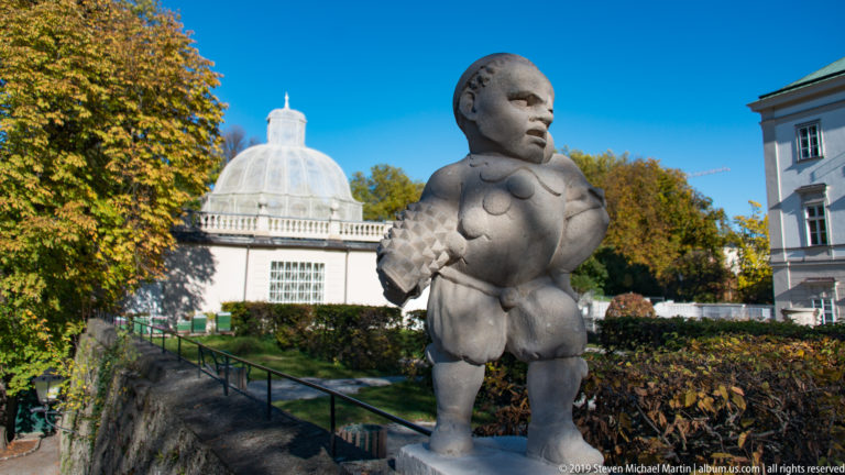 Stone dwarf statue in period costume standing in Mirabell Gardens, with a glass-domed pavilion and autumn plantings behind.