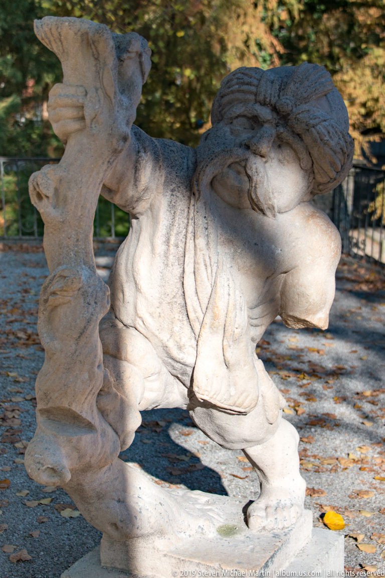 Bent stone dwarf carrying a large burden across the back while gripping a vertical support or staff.