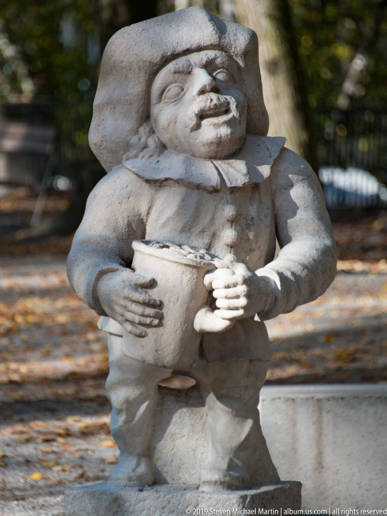 Front-facing stone dwarf in broad hat and ruff collar holding a large handled vessel against the abdomen.